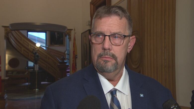 A man in a suit and glasses stands in a hallway talking to reporterss.