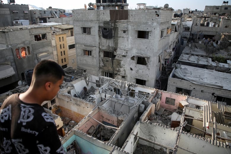 A man on a roof in Rafah looks down at a series of damaged rooms with no roof. 