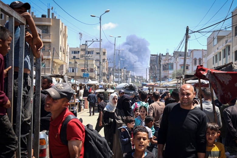 People walk along a busy urban street as black smoke billows in the background.