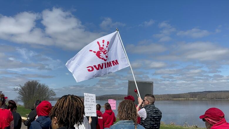 A crowd of people wearing red clothing walk down a street next to a wide river. One marcher holds up a white flag with a red handprint and letter MMIWG on it.