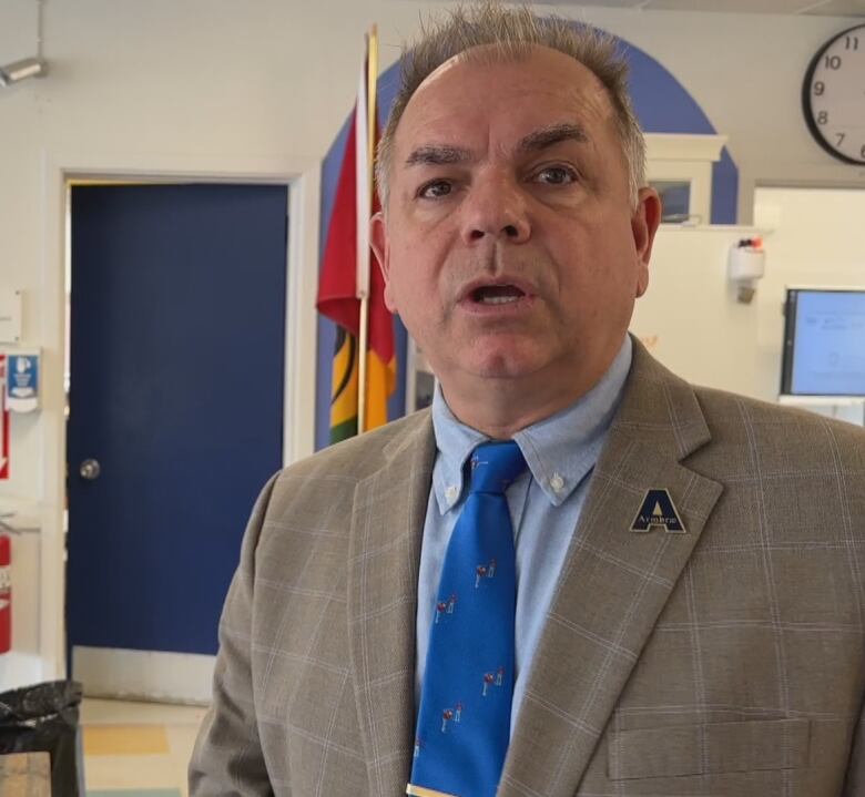 A man wears a gray blazer and a blue tie and a pin bearing the letter A. 