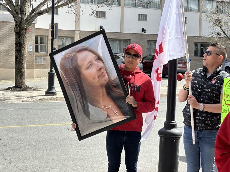 A man wearing a red sweater and red baseball cap stands on a street holding up an enlarged photo of a woman's face.