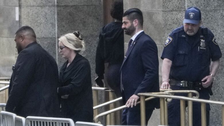 A woman wearing glasses with her hair tied up is shown walking behind metal barriers outside a building, with two men on either side of her.