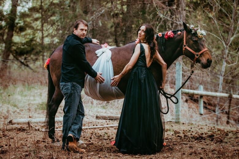 Man and woman wearing nice clothes have hands on the belly of a pregnant brown horse who's stomach is wrapped in transparent white fabric. 