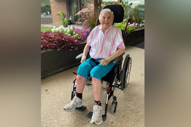 Woman with grey hair, pink and white shirt and blue shorts sits outside in a wheelchair. 