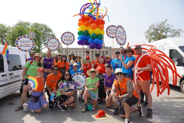 Photo of many people in multi-coloured CBC Pride shirts, carrying balloons and smiling