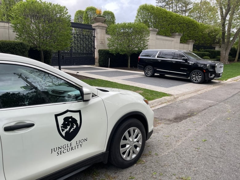 A white crossover SUV and a large black SUV are seen parked outside black gates.