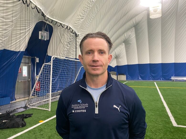 A man wearing a blue jacket stands on a green turf field in font of a soccer net under a white tent.