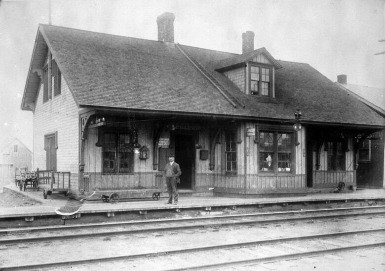 A black and white photo of a wooden station house with decorative mouldings beneath the overhang. A man in a suit and hat with his hands in his pockets on the platform. Two sets of empty tracks in the foregound.