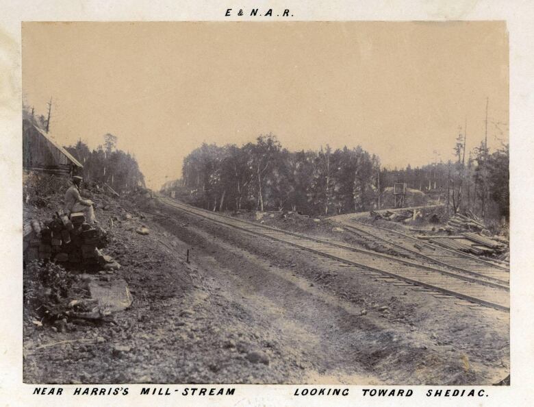 A man sitting on a pile of timbers in the left foreground. A log cabin behind him. Train tracks stretching into the distance from left to right diagonally through a cut of trees. Written in the margin of the old photo: E.&N.A.R. near Harris's Mill-stream looking toward Shediac.