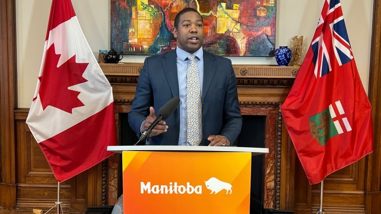 A man is a suit is pictured speaking at a podium that says 'Manitoba,' in between a flag of Manitoba and Canada.