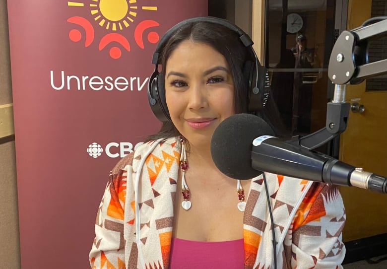 A dark-haired woman, wearing a headset, sits in front of a microphone with a banner that reads 