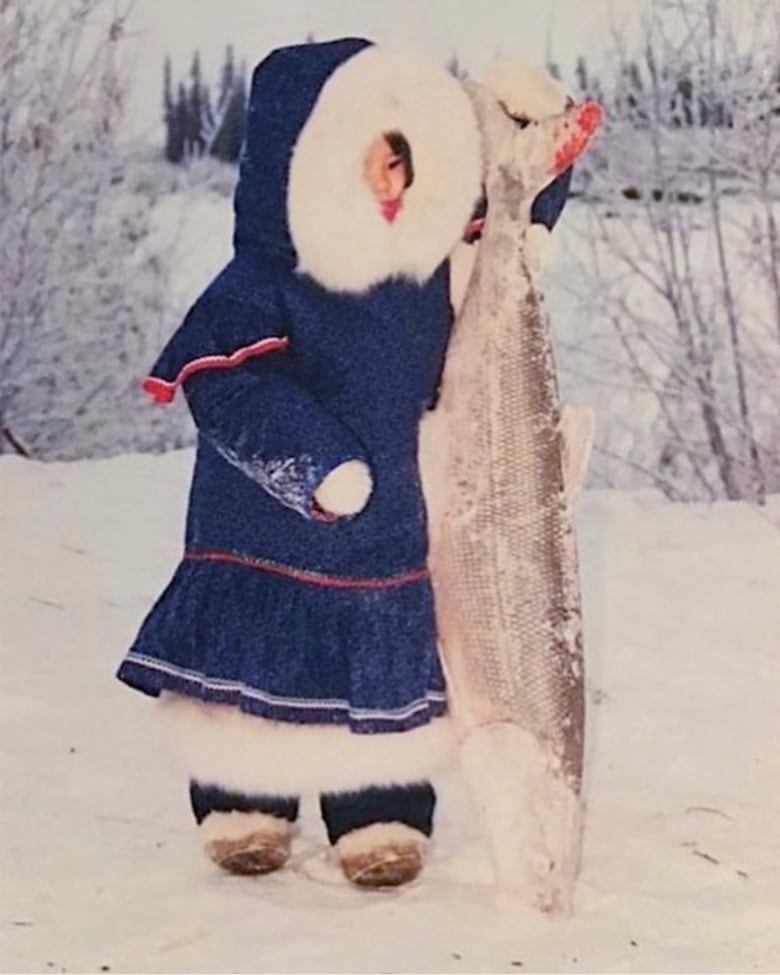 A young child in Indigenous winter clothing holds a large fish.