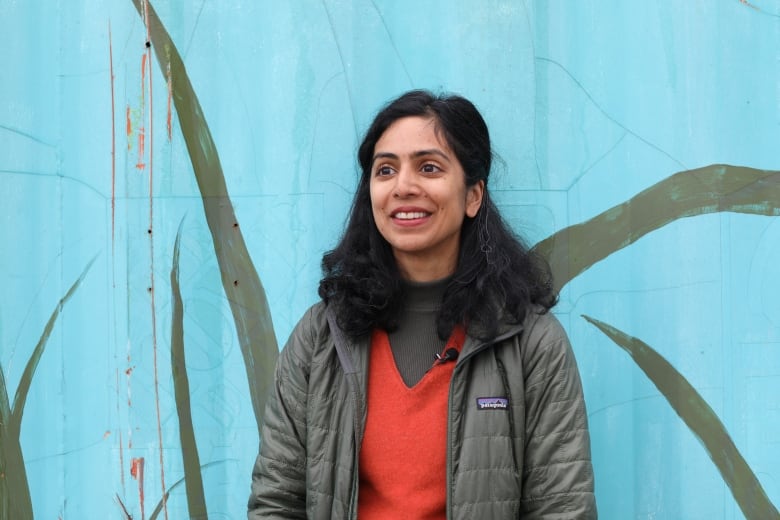 A young adult woman with long black hair, wearing a green-grey spring jacket over an orange shirts, stands in front of a blue wall.