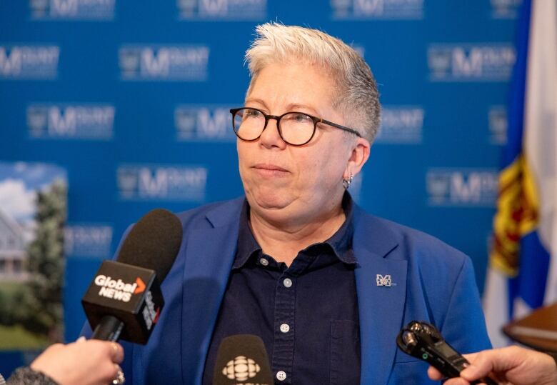 A person with glasses and short hair stands in front of microphones.