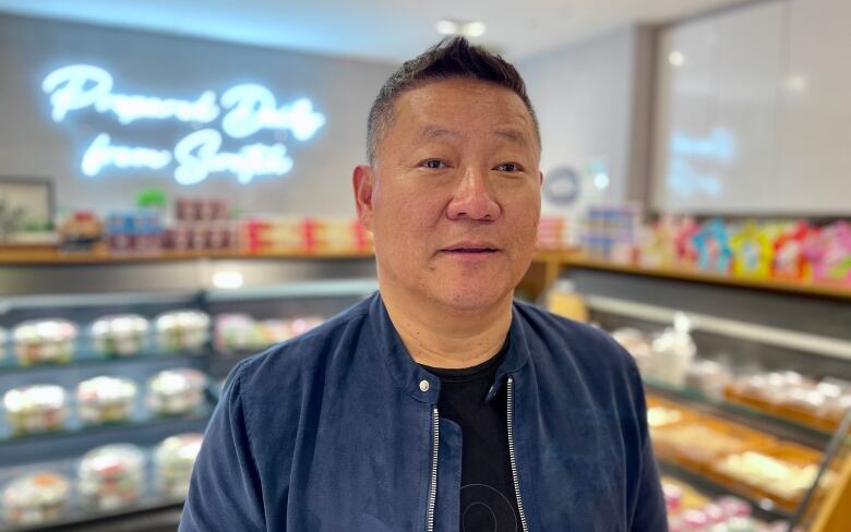 An East Asian man in a blue jacket stands inside a food retailer.