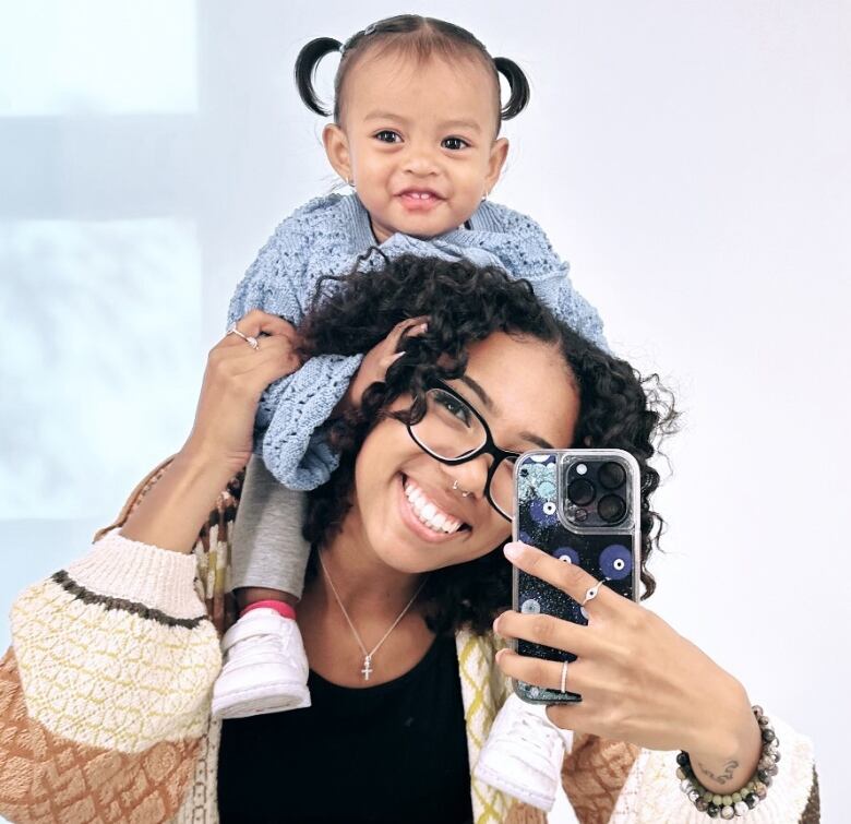 A young woman poses in a mirror with a baby on her shoulder. She is taking a selfie in the mirror with an Iphone. 