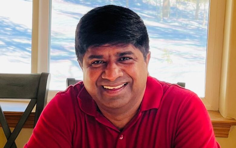 A South Asian man in a red shirt sits in front of a lakeview window.