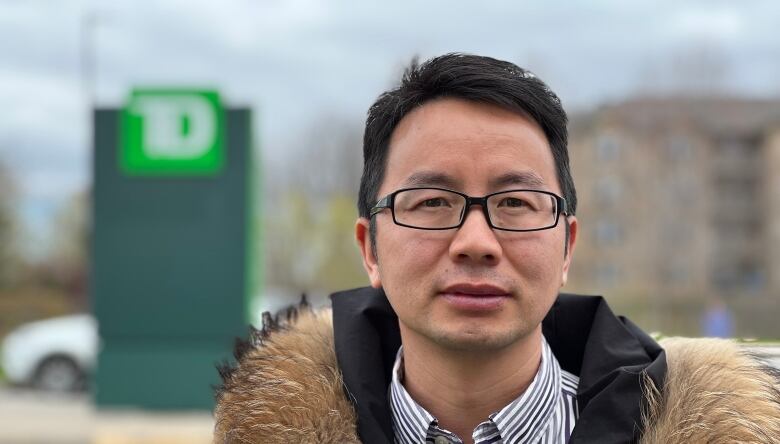 An East Asian man wearing glasses and a coat with fur trim stands in a parking lot in front of a blurred TD Bank logo.