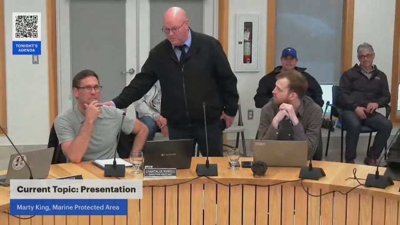 a man stands behind two sitting at a testimony table in a council chamber. 