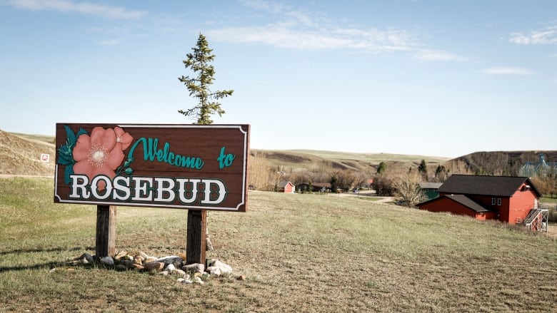 A welcome sign to the hamlet of Rosebud, Alberta, is seen in the foreground, with rolling hills and a red barn in the background.