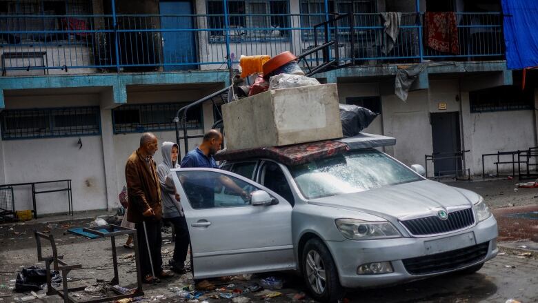 Three people are shown at the side of a sedan which has items loaded atop the vehicle's roof. Debris litters the ground near the structure where the vehicle is parked.