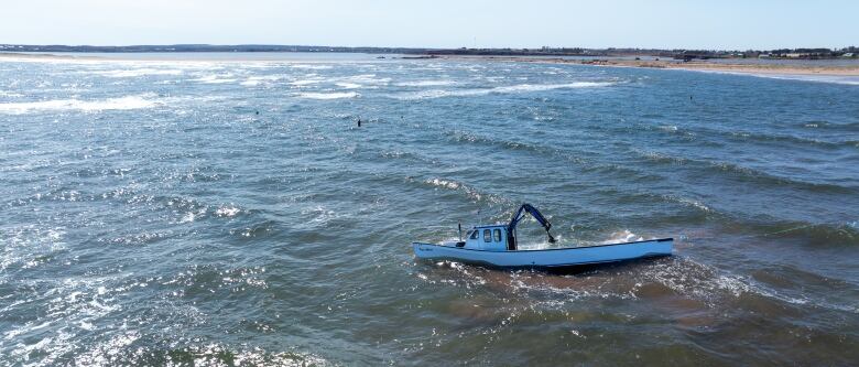 A drone view of a half-submerged white and blue fishing boat near a sandbar, being hit by waves. 