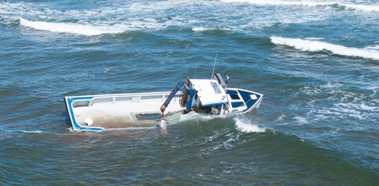 A drone view of a half-submerged white and blue fishing boat near a sandbar, being hit by waves. 