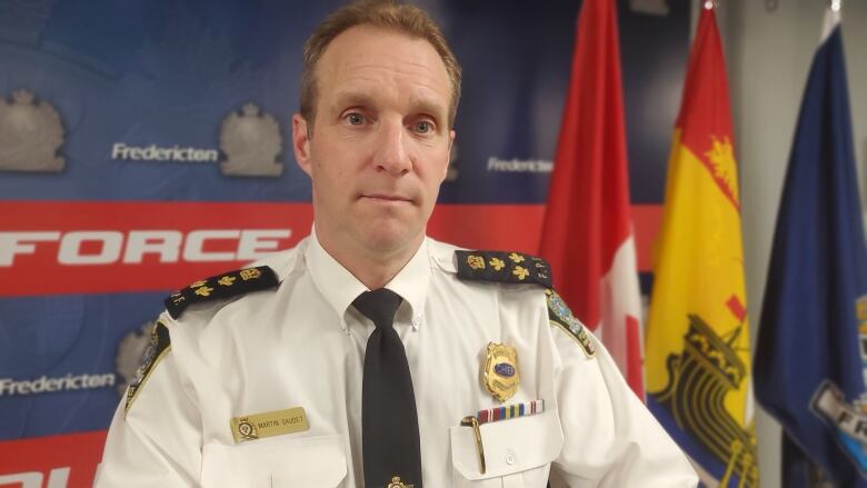 A man with brown hair wearing a white police uniform stands in front of a wall with the Fredericton Police Force logo on it and a Canadian flag.