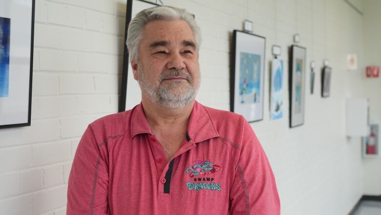 A grey haired and bearded man wearing a salmon coloured shirt poses indoors in front of a white brick wall with paintings hanging in the background. 