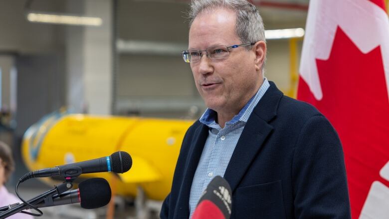 A man wearing a blazer and a blue oxford shirt stands in front of several microphones. In the background, a yellow slocum glider can be seen.  