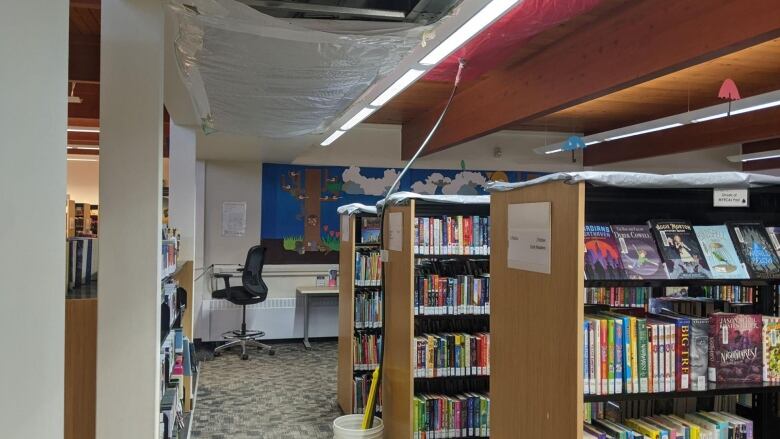 The inside of a library is shown with roof panels removed and tarps covering the hole in the roof as well as lining the bookshelves below.