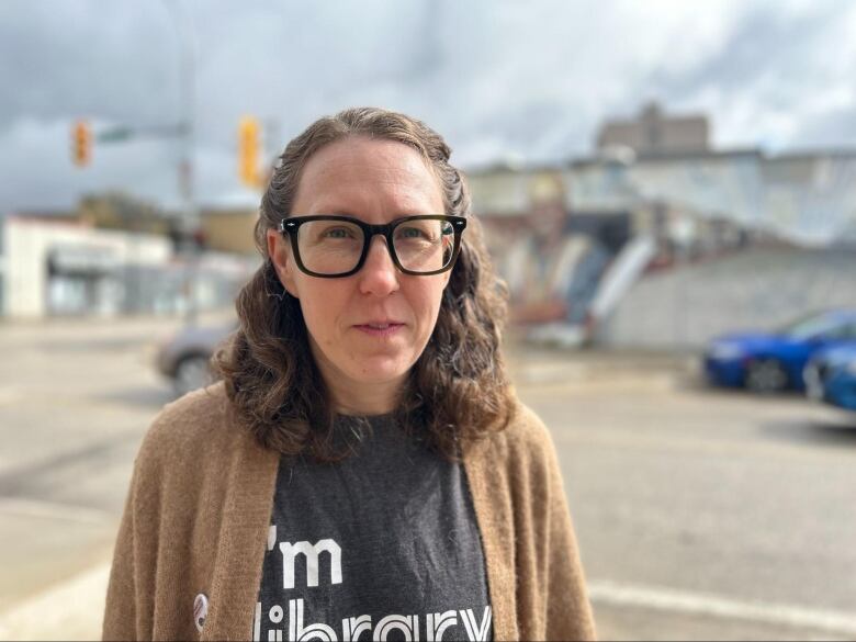 A woman wearing dark-rimmed glasses and a light brown cardigan over top a dark grey T-shirt stands at an intersection.