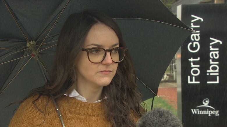 A woman wearing dark-rimmed glasses and a mustard-coloured sweater and holding a black umbrella 