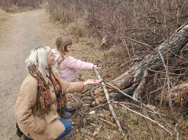 A woman in brown coat leans next to a little girl with blond hair as they hold out their hand and feed a bird on a path in a park.