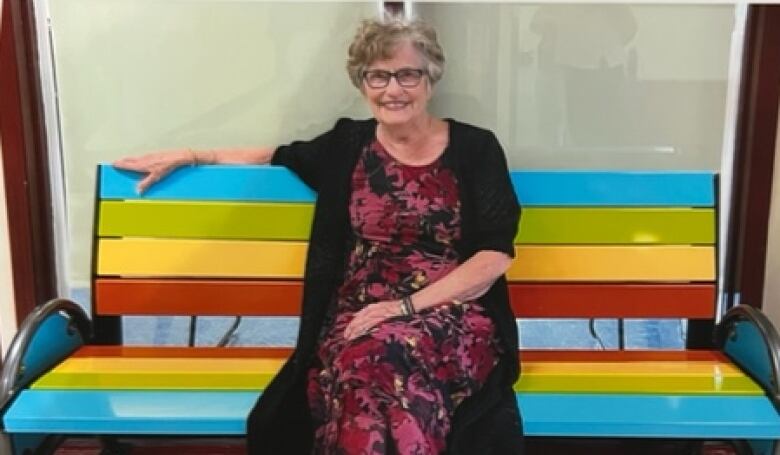 A woman sits on a rainbow coloured bench at a school. 