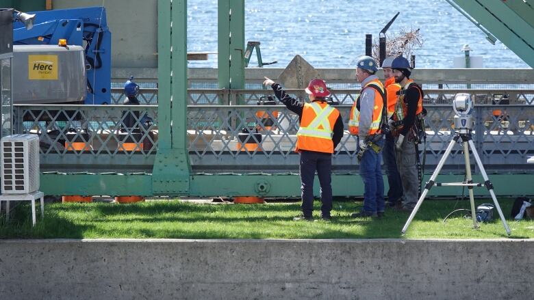 Four people wearing bright orange vests and hard hats are shown standing next to a green, metal bridge.