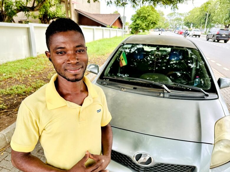 A young man wearing a yellow polo shirt stands in front of a silver car with a small Ghana flag visible behind the windshield. 