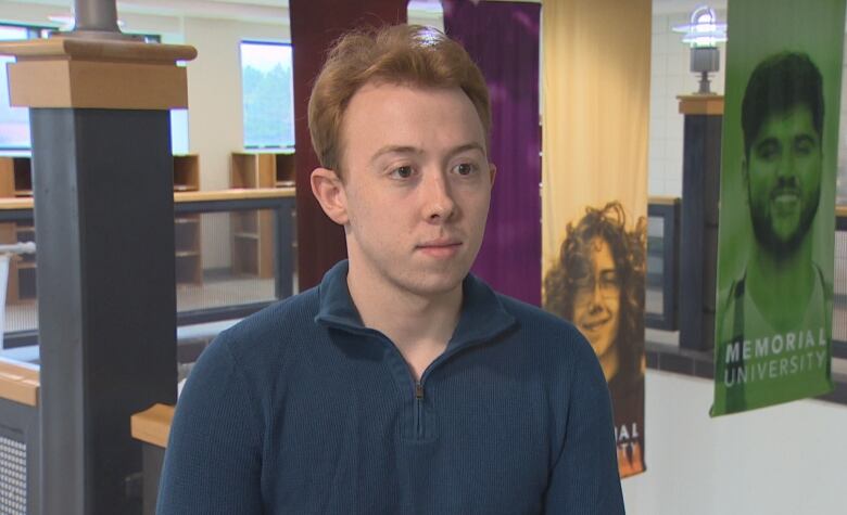 A man with red hair wearing a blue sweater stands inside a university.