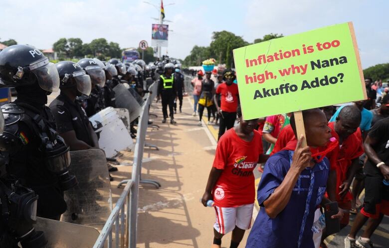 Crowds of people holding signs march in the street next to a metal barricade lined by uniformed police officers wearing helmets and face shields.