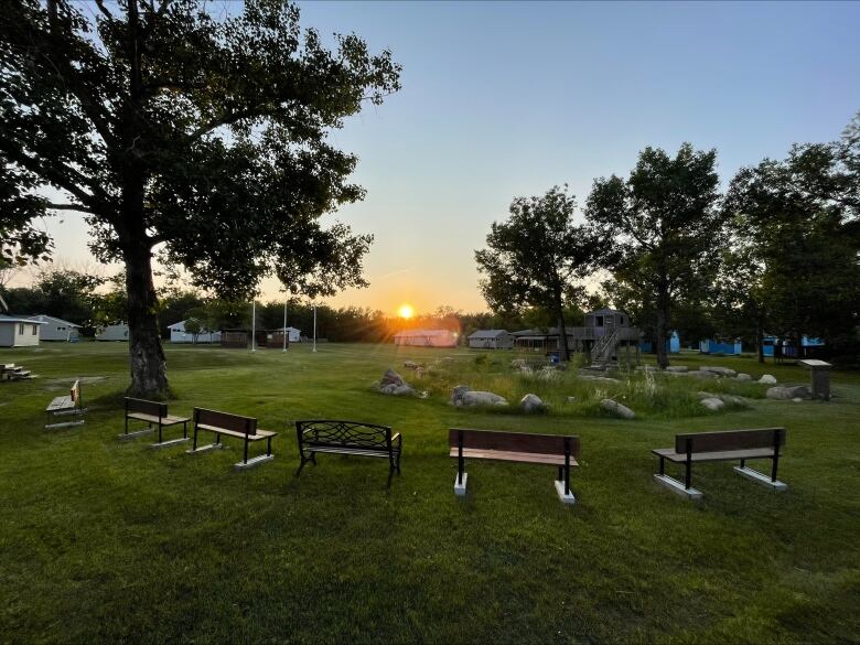 A sun sets over a grassy field with benches and trees.