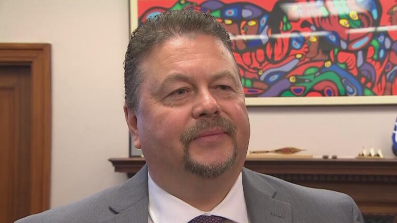 A man with short grey hair and a goatee wearing a grey blazer and purple tie stands in an office.