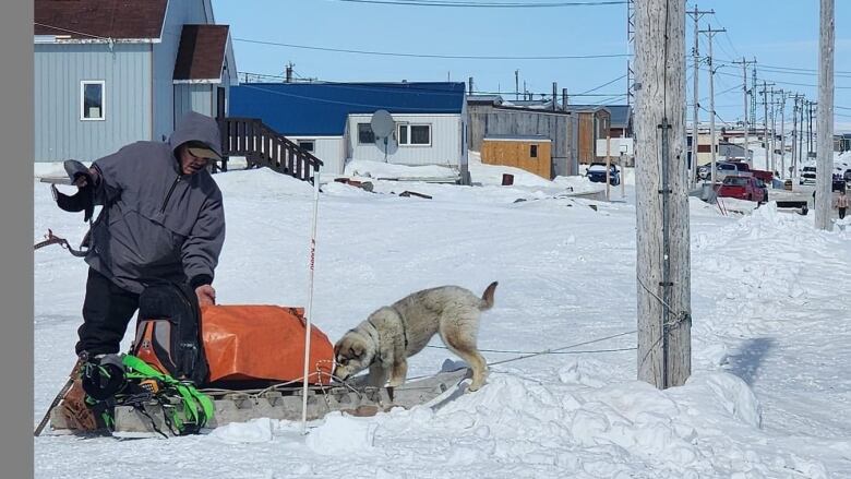 A man in a community with snow on the ground and a wooden sled with a dog 