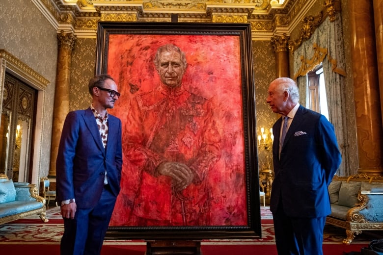 Two men in blue suits stand in front of a red-hued portrait of King Charles III.