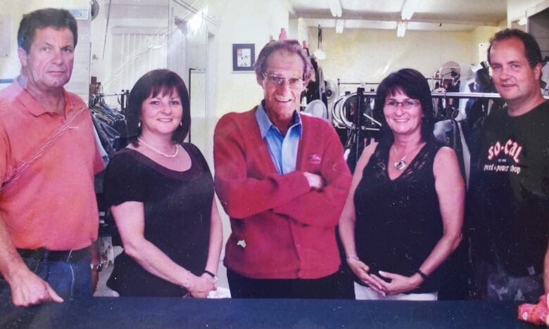 Five people stand in a row. In the centre, in a red sweater, is a smiling older man with his arms folded.