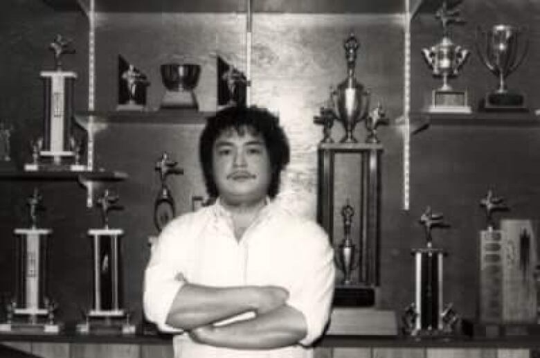 A black-haired man with a thin mustache and a white shirt stands with his arms crossed, with trophies seen behind him.