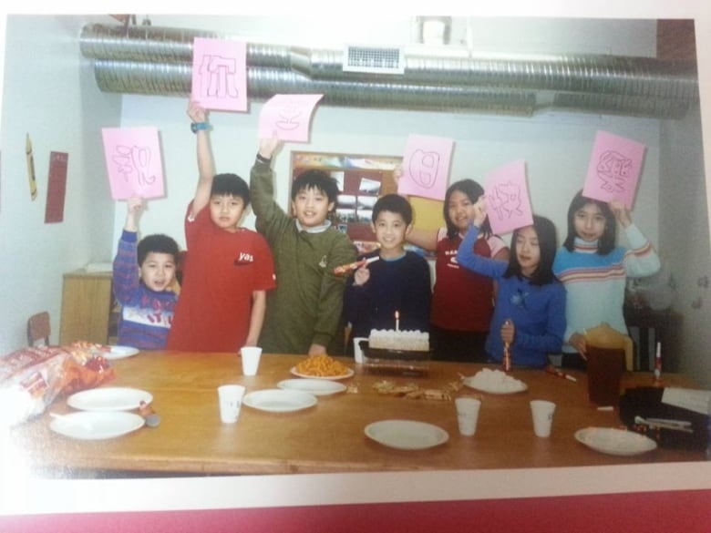 Seven children holding up pink papers stand in line behind a cake at a kitchen table.