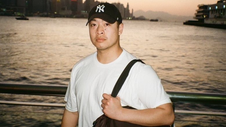 A man wearing a black ball cap and a white T-shirt can be seen in front of a body of water, a bag slung over his shoulder.