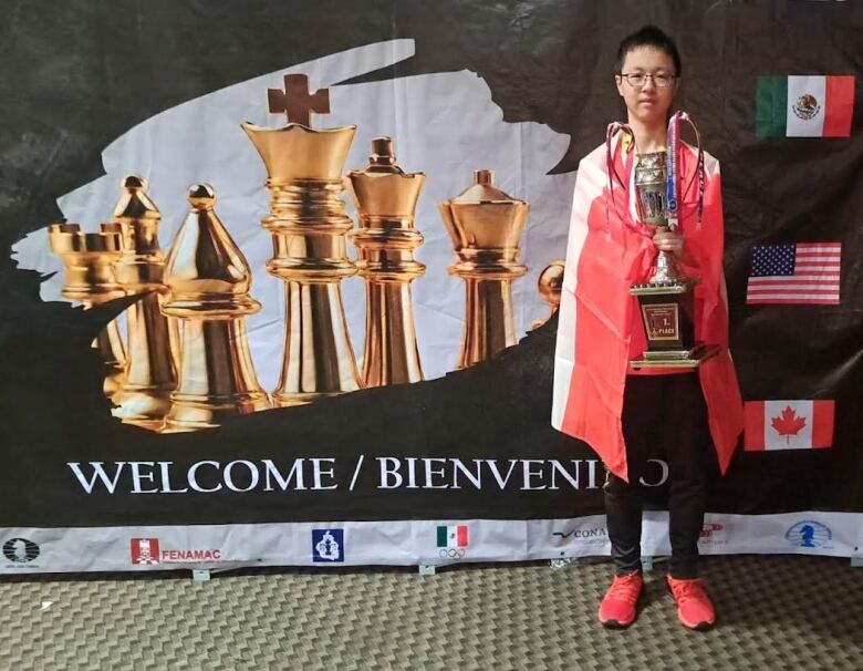 A boy stands in front of a banner with chess pieces on it, holding a trophy.