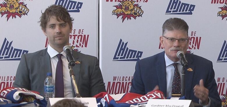 Two mean sit behind a table decorated with graphics of the Moncton Wildcats logo.